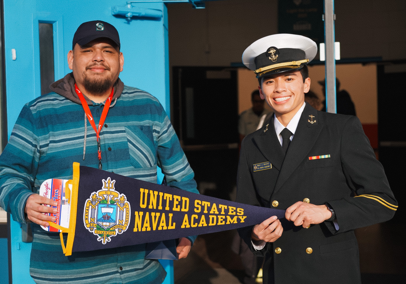 USNA Midshipman and a man in blue sweatshirt holding a USNA flag.